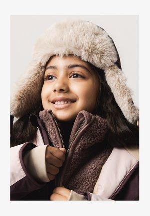 Lächendes Kind mit pelziger Wintermütze und Reißverschlussjacke, hält den Reißverschluss der Jacke mit beiden Händen.
