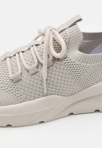 Beige knit sneaker with a textured upper, elastic laces, and a chunky rubber sole, featuring a pull tab at the heel for easy wear.