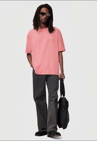T-shirt oversized rosa claro com logótipo branco, combinado com jeans cinza escuro de perna reta e sapatos pretos. Segurando uma mala preta.