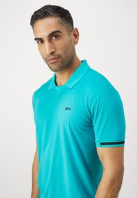Turkizna polo majica iz teksturirane tkanine, s crnim "BOSS" logotipom na prsih i crnim prugama na rukavima.