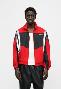 Άνδρας που φοράει αθλητικό μπουφάν Adidas κόκκινο και μαύρο πάνω από λευκή φανέλα, συνδυασμένο με μαύρο παντελόνι, σε απλό λευκό φόντο.