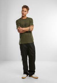 Olīvu zaļš T-krekls ar mazu melnu logotipu uz krūts, apvienots ar melniem kabatas biksēm ar vairākiem kabatām un rāvējslēdzējiem, valkāts ar baltām sporta apavām.