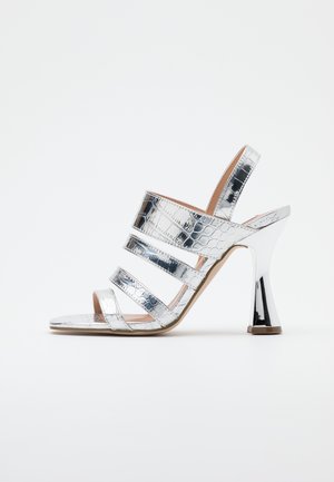 Srebrne sandały na wysokim obcasie z teksturą krokodylej skóry, cztery poziome paski i szeroki, zwężający się obcas na białym tle.