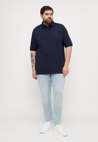 Marineblauwe polo met korte mouwen, kraag en een klein logo. Gedirigeerd met lichtblauwe spijkerbroek en witte sneakers. Eenvoudig ontwerp.