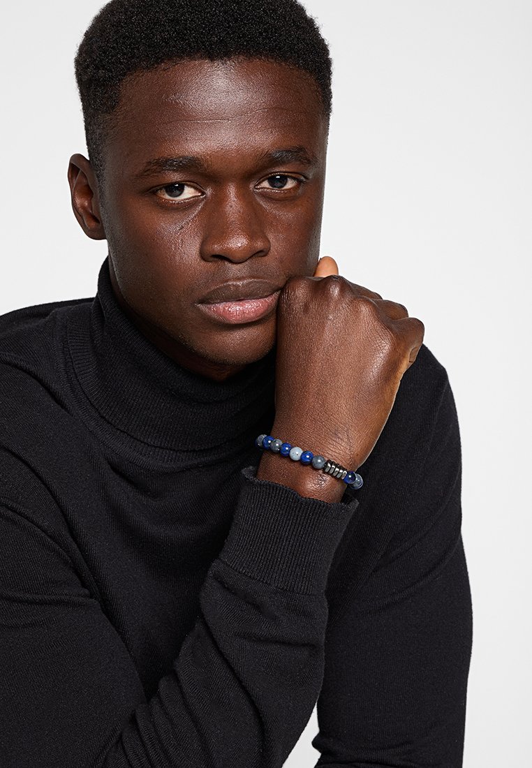 Maglione a collo alto nero, modello che indossa un braccialetto di perline multicolore con perline blu e nere e accenti metallici.