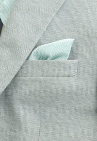 Hellgrauer, strukturierter Blazer mit einem hellblauen, gemusterten Einstecktuch, das in die Brusttasche gesteckt ist.