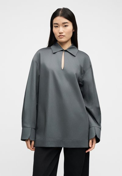 Γυναίκα που φοράει φαρδιά γκρι μπλούζα με μακριά μανίκια, φαρδιές μανσέτες και μικρό άνοιγμα μπροστά, συνδυασμένη με μαύρο παντελόνι, στέκεται μπροστά σε απλό φόντο.