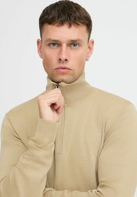 Beige, ribbet zip-up genser med høy krage, laget av mykt stoff. Har lange ermer og en slank passform.