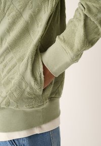 Felpa verde con trama testurizzata caratterizzata da motivi in rilievo, polsini a coste e una tasca a marsupio. Indossata sopra una camicia bianca e jeans.