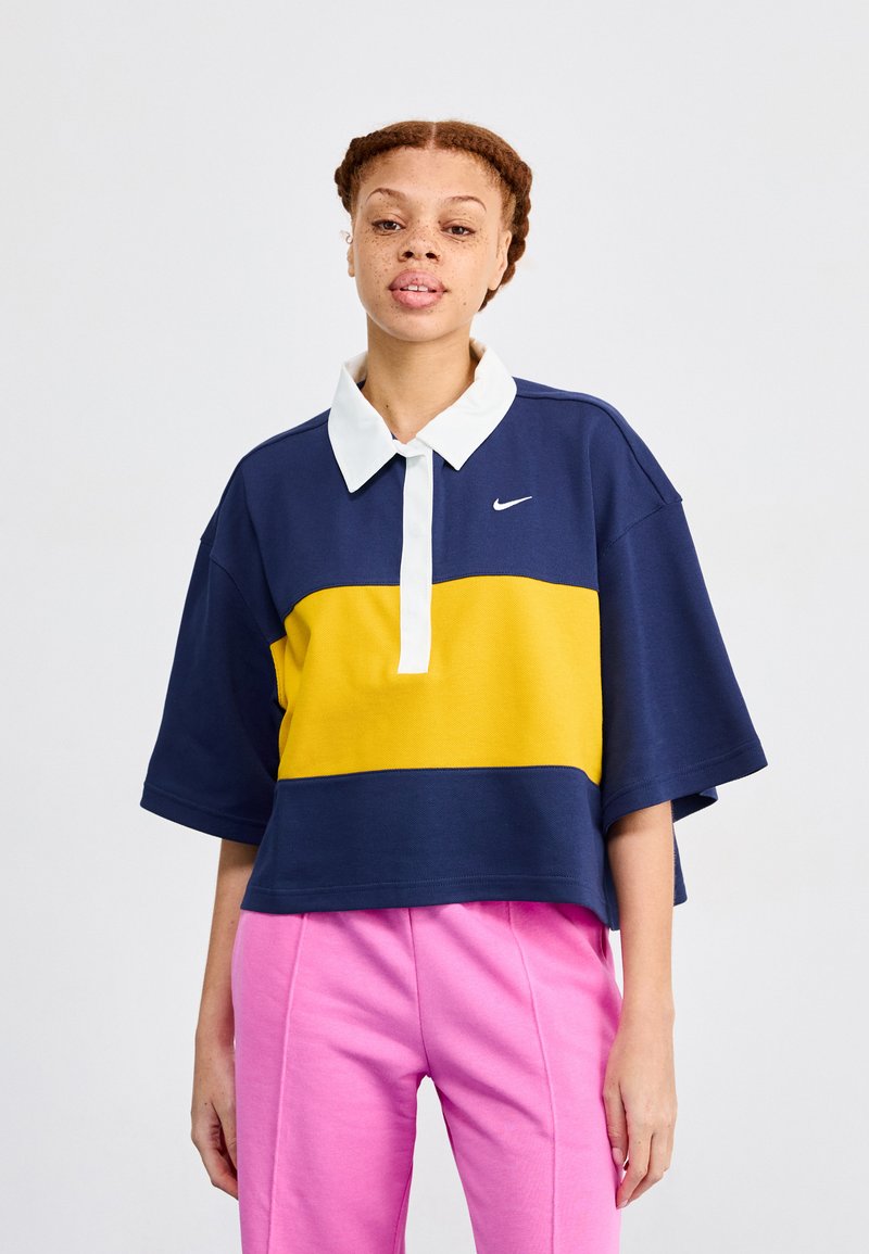 Navy blue, yellow, and white polo-style top with short sleeves, featuring a collar and a Nike logo. Paired with pink sweatpants.