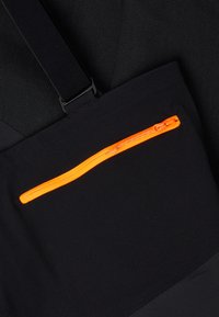 Svart stoff med en jevn tekstur har en liten, horisontal oransje glidelåslomme. Justerbar svart stropp i bakgrunnen.