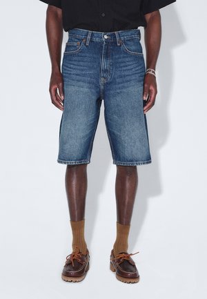 Mężczyzna ubrany w średnioniebieskie szorty dżinsowe, połączone z brązowymi skarpetkami i ciemnobrązowymi skórzanymi butami na sznurowadła, stojący na jednolitym tle.