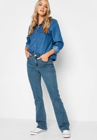 Niebieska koszula dżinsowa z dwiema kieszeniami na piersi, zestawiona z jasnoniebieskimi dzwonami i białymi trampkami, na jednolitym białym tle.