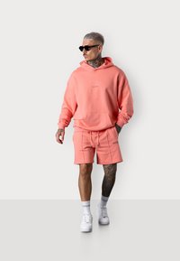 Hoddie pêche avec poche avant et logo, associé à un short assorti. Des baskets blanches et des lunettes de soleil complètent le look. Tissu lisse et décontracté.