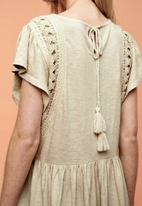 Beiges Kleid mit kurzen, gerafften Ärmeln. Verfügt über Häkelverzierungen an den Schultern und eine Quastenbindung am Rücken. Weicher, strukturierter Stoff.