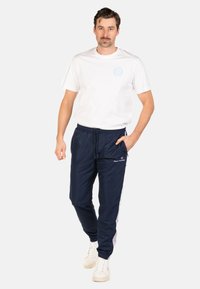 Herenoutfit met een plain wit T-shirt en marineblauwe trainingsbroek met elastische tailleband, zijzakken en logo-accenten. Witte sneakers.
