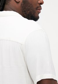 Short-sleeve shirt in white fabric features a smooth texture, a yoke design at the back, and subtle stitching details.