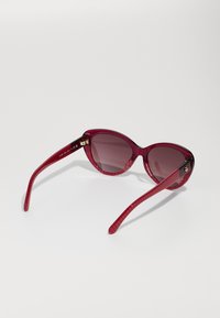 Γυαλιά ηλίου cat-eye με σκούρο κόκκινο σκελετό και φακούς με κλίση χρώματος. Οι τεχνικές κορώνες διαθέτουν χρυσές λεπτομέρειες.
