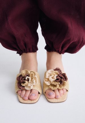 Pies que llevan sandalias beige tejidas tipo slip-on con grandes flores de crochet, combinadas con pantalones elásticos burdeos hasta el tobillo sobre un suelo blanco.