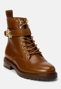 Bota de tornozelo em pele castanha com acessórios dourados, design de atacadores, faixa lateral com logotipo e sola volumosa para maior tração.
