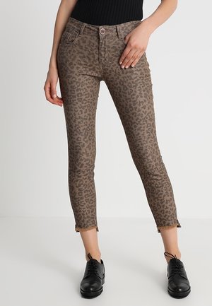 Kobieta w krótkich brązowych wąskich spodniach w panterkę z postrzępionymi zakończeniami i czarnych sznurowanych butach, ręce spoczywają blisko kieszeni.