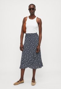 Débardeur blanc avec une jupe midi florale bleu marine, ornée de petites fleurs blanches. Un sac à main marron et des chaussures slip-on beiges complètent le look.
