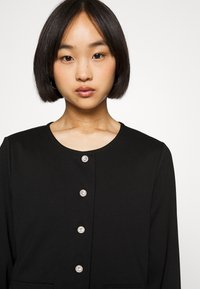 Fekete dzseki kerek nyakkivágással, hosszú ujjal és hat elülső gombbal, fényes fémes kivitelben. Egyszerű és klasszikus dizájn.