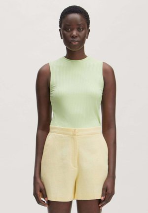 Femme aux cheveux courts porte un haut côtelé sans manches vert clair et un short taille haute jaune pâle, debout devant un fond clair uni.