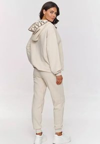 Beżowa bluza z kapturem z logo, w komplecie z beżowymi dresami. Miękka tkanina, luźny krój i ściągacze na rękawach. Białe sneakersy.