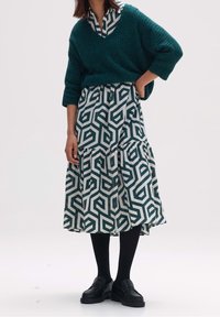 Stickad grön tröja med V-ringning, parat med en lager-på-lager kjol med ett geometriskt mönster i mörkgrön och vit. Svarta skor och strumpbyxor.