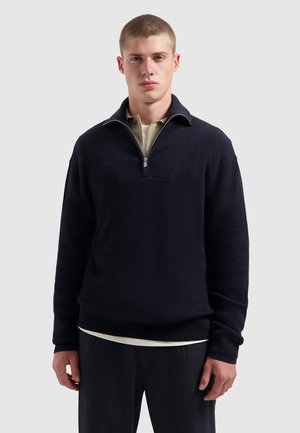 Marineblauw geribbeld sweater met een kwart rits en een hoge kraag, voorzien van een contrasterende binnenlaag die zichtbaar is bij de hals. Heeft een losse pasvorm.