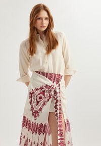 Chemise beige clair avec un design à bouton. Associée à une jupe enroulée blanche ornée de motifs géométriques et floraux rouges, nouée à la taille.