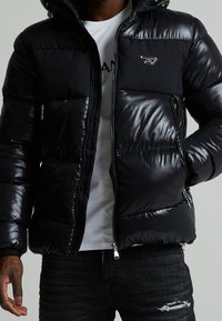 Schwarze Puffjacke mit glänzendem Finish, kontrastiert durch matte Stoffpaneele. Verfügt über eine Kapuze, Reißverschlusstaschen und ein Logo-Patch auf der Brust.
