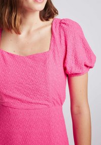 Robe texturée fuchsia avec décolleté carré et manches bouffantes, fabriquée dans un matériau léger avec une finition froissée.