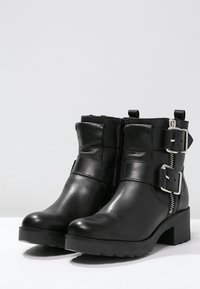 Svarta ankelstövlar i blank läder med en kraftig klack, utrustade med en dragkedja och två dekorativa spännen på sidan.