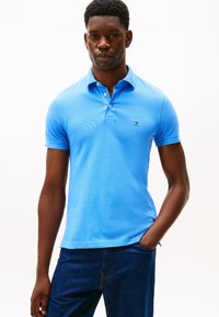 Lys blå poloshirt med korte ermer og en treknapps krage. Har en liten logo på venstre bryst. Kombinert med mørke denimbukser.