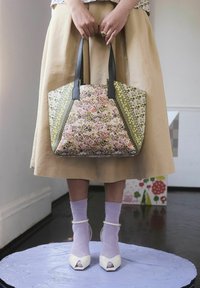 Kobieta stojąca na okrągłej lawendowej platformie, trzymająca dużą torbę na ramię w kwiatowy wzór, ubrana w beżową spódnicę, lawendowe skarpety i białe szpilki.