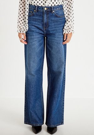 Vrouw draagt blauw, hooggetailleerde, wijd uitlopende jeans, zwarte puntige schoenen en een witte blouse met zwarte stippen en lange mouwen.
