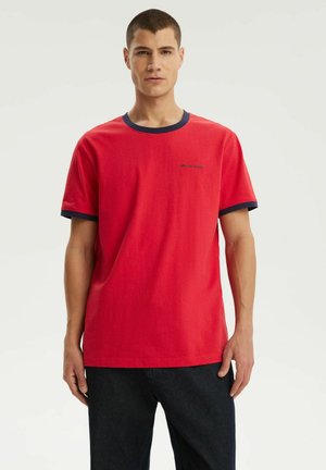 Młody mężczyzna stojący, ubrany w czerwony t-shirt z krótkim rękawem z ciemnoniebieskim kołnierzem i lamówkami na rękawach, w połączeniu z czarnymi spodniami na jednolitym tle.