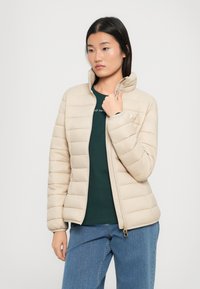Vrouw met steil zwart haar die een beige gewatteerde jas, een donkergroen shirt en blauwe spijkerbroek draagt, terwijl ze met één hand de kraag van de jas vasthoudt.