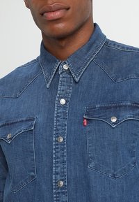 Tummansininen farkkupaita, jossa on napillinen kaulus, kaksi rintataskua napilla, ja hopeanväriset nepparit. Sauman yksityiskohtia mukana.