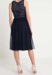 Robe de cocktail bleu marine avec un corsage perlé et une jupe en tulle transparente. Présente un design sans manches et arrive jusqu'à mi-mollet.