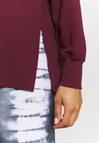 Bordeauxrotes Langarmshirt mit seitlichem Schlitz. Gerippte Bündchen und Saum. Getragen über grauen Leggings mit Batikmuster. Glatte Textur.