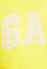 Indumento giallo con grandi lettere ricamate "G" e "A" in outline bianco. Il tessuto appare liscio con una texture morbida.