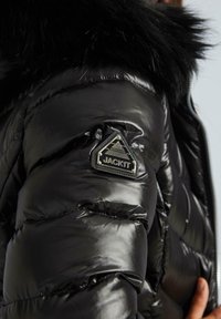 Melnā pūkainā jaka ar spīdīgu, quiltētu tekstūru, ar melnu kažokādu kapuci un trīsstūrveida uzlīmi ar "JACKIT" logotipu uz piedurknes.