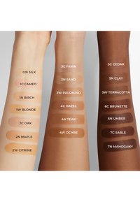 Väri näyteitä meikkivoiteesta eri beige- ja ruskeasävyissä iholla, joissa on väri-nimet ja koodit, esittäen sileitä, mattaisia viimeistelyjä.