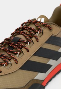 Καφέ αθλητικό παπούτσι με υφασμένο επάνω μέρος, μαύρα και πορτοκαλί κορδόνια, τρεις μαύρες ρίγες και κόκκινη λεπτομέρεια στη σόλα.