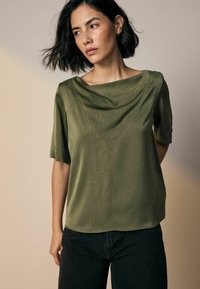 Olijfgroene blouse met een gedrapeerde colkraag, korte mouwen en een textuurafwerking, gecombineerd met donkere jeans. Eenvoudige, losse pasvorm.