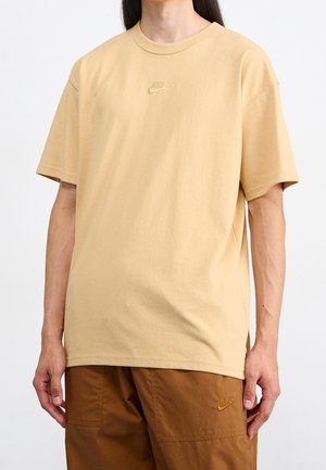 Persoon draagt een beige Nike T-shirt en bruine Nike broek, staat met ontspannen armen tegen een effen witte achtergrond.