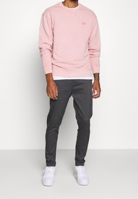 Světlá růžová mikina s žebrovanými manžetami, doplněná tmavě šedými kalhotami s proužky a bílými teniskami. Model stojí na neutrálním pozadí.